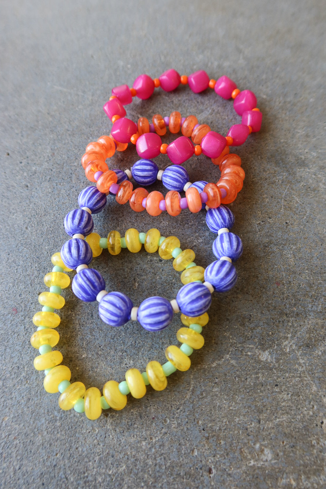 Four beaded stretch bracelets in pink, orange, purple, and yellow, arranged on a textured gray surface