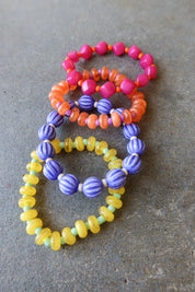 Four beaded stretch bracelets in pink, orange, purple, and yellow, arranged on a textured gray surface