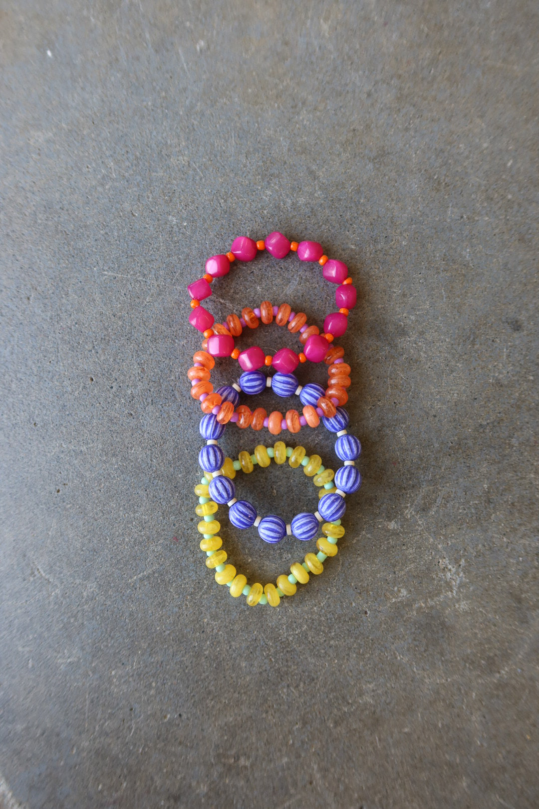 Three beaded stretch rings in pink, orange, and yellow with purple accents, arranged vertically on a textured gray surface