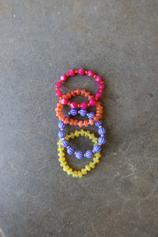 Three beaded stretch rings in pink, orange, and yellow with purple accents, arranged vertically on a textured gray surface