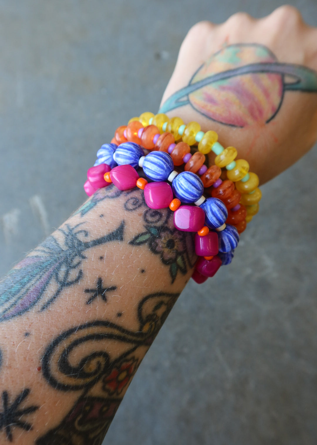 A close-up of a tattooed wrist wearing multiple colorful beaded bracelets, including yellow, orange, blue, pink, and purple beads, against a gray concrete background