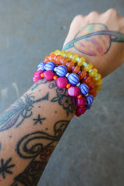 A close-up of a tattooed wrist wearing multiple colorful beaded bracelets, including yellow, orange, blue, pink, and purple beads, against a gray concrete background