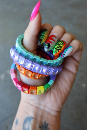 A close-up of a hand holding a stack of chunky, colorful beaded bracelets with rhinestones. The bracelets feature vibrant rainbow colors including pink, orange, yellow, green, blue, and purple, with some featuring fish designs on the front