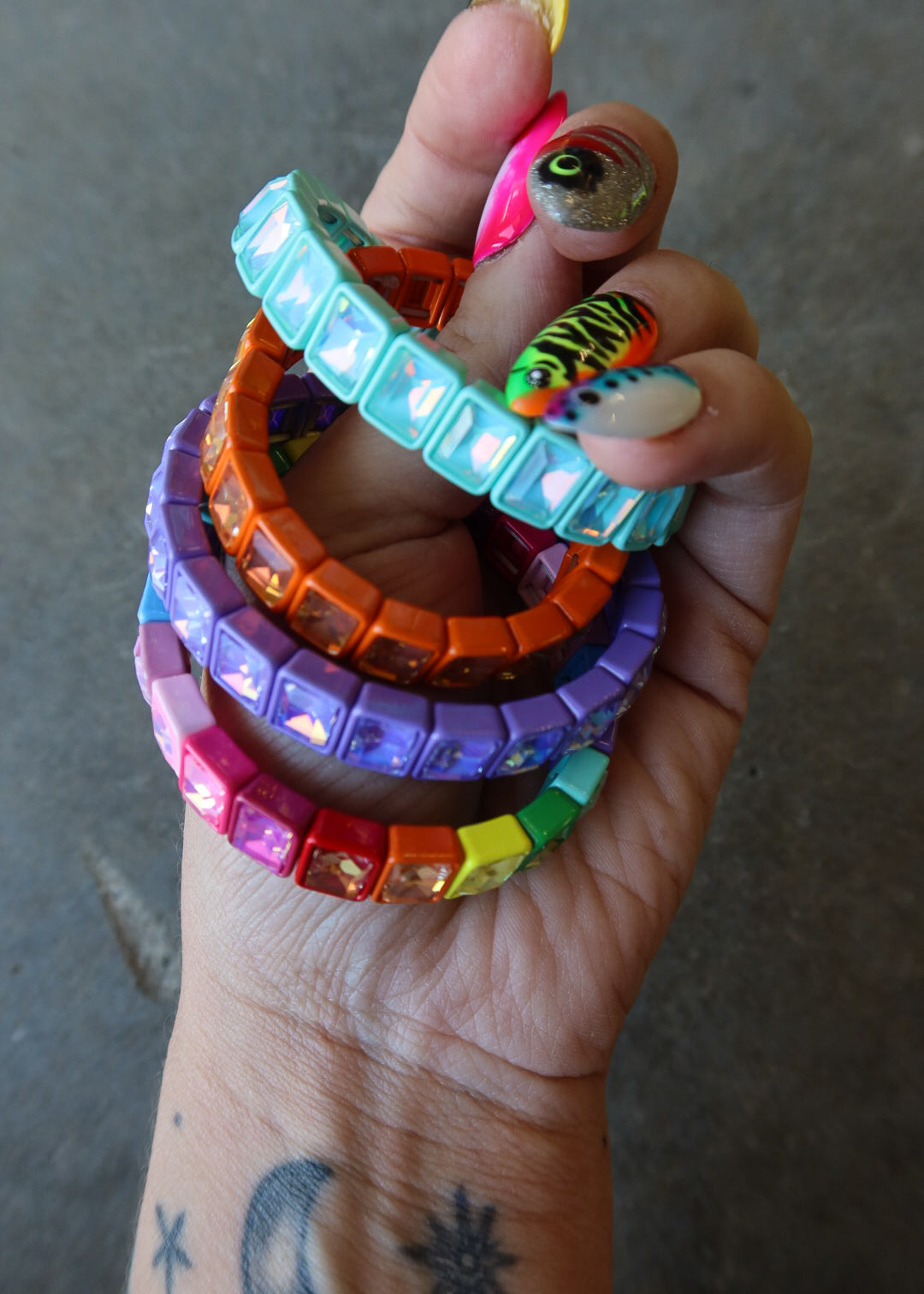 A hand holding a stack of colorful rhinestone-embellished bracelets in turquoise, orange, purple, pink, yellow, green, and rainbow colors, against a gray concrete background
