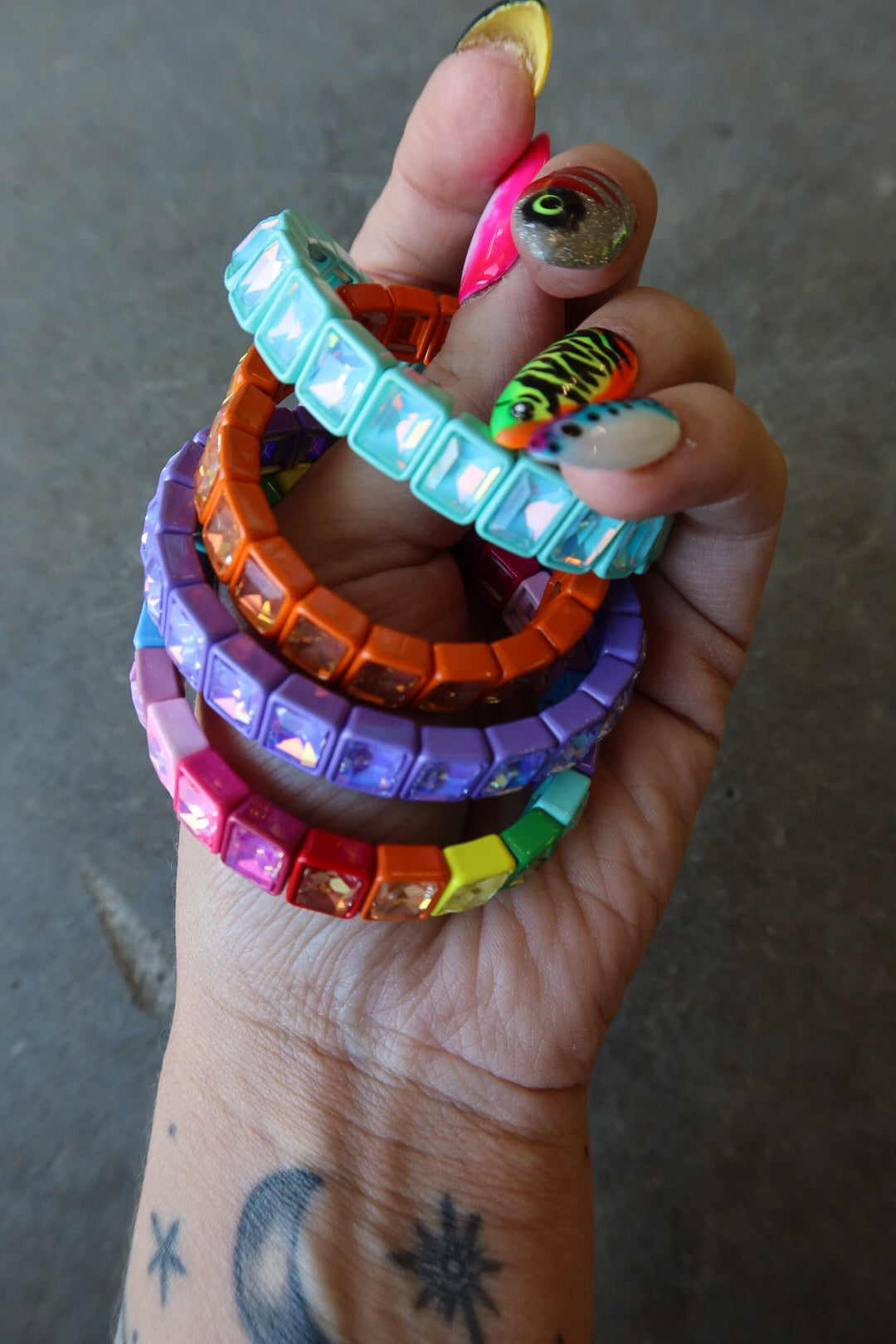 A hand holding a stack of colorful rhinestone-embellished bracelets in turquoise, orange, purple, pink, yellow, green, and rainbow colors, against a gray concrete background