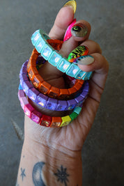 A hand holding a stack of colorful rhinestone-embellished bracelets in turquoise, orange, purple, pink, yellow, green, and rainbow colors, against a gray concrete background
