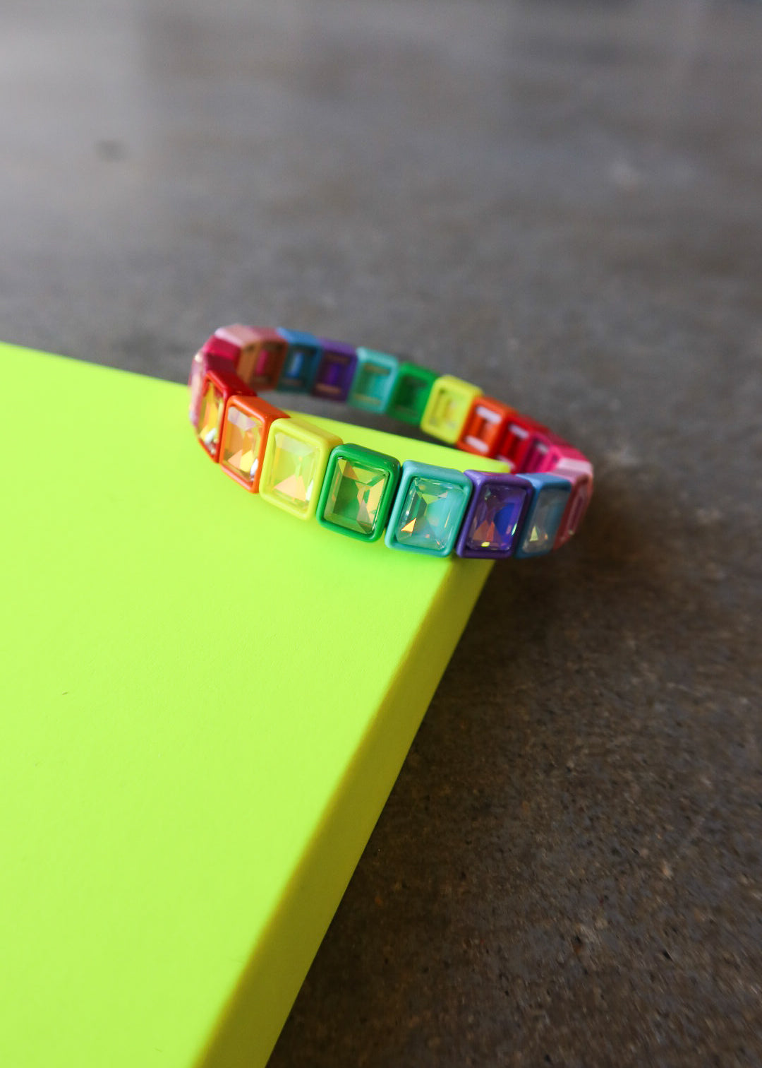 A colorful stretch bracelet with square rainbow-colored rhinestones resting on a bright neon yellow-green surface against a dark gray concrete floor.