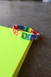A colorful stretch bracelet with square rainbow-colored rhinestones resting on a bright neon yellow-green surface against a dark gray concrete floor.