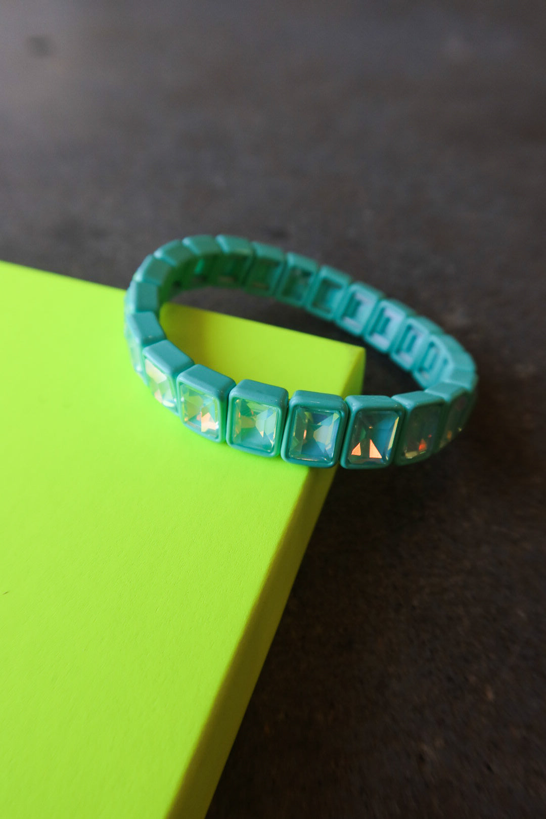 A turquoise beaded bracelet with square iridescent rhinestones, resting on a bright neon yellow-green rectangular block against a dark textured background
