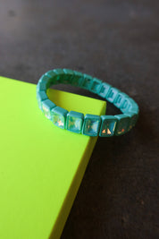A turquoise beaded bracelet with square iridescent rhinestones, resting on a bright neon yellow-green rectangular block against a dark textured background