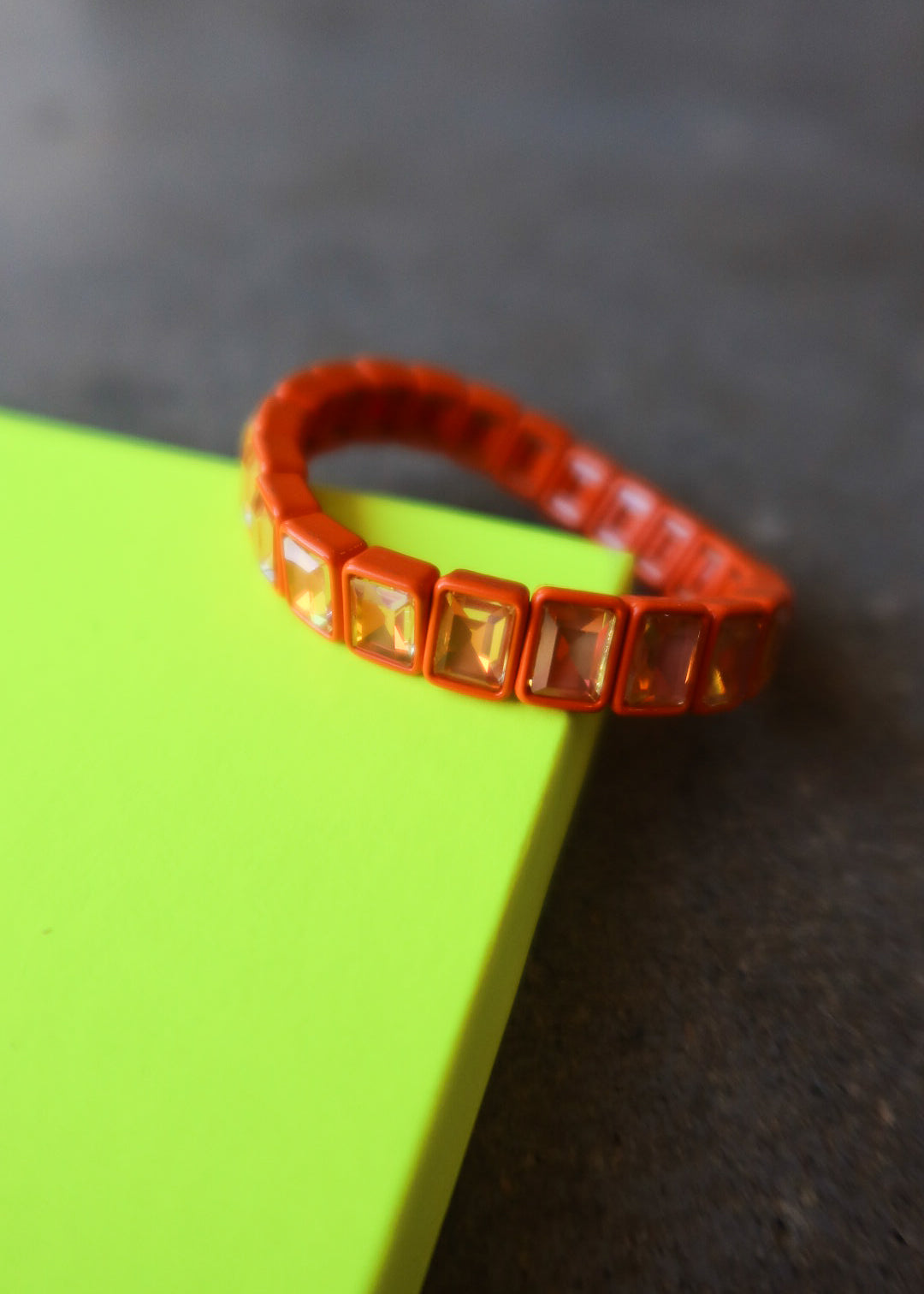 An orange beaded bracelet with square iridescent rhinestones, resting on a bright neon yellow-green rectangular block against a dark textured background