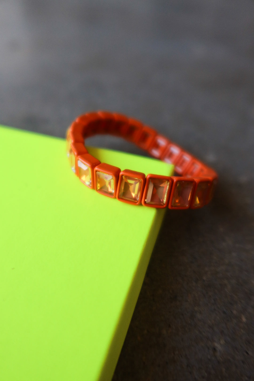 An orange beaded bracelet with square iridescent rhinestones, resting on a bright neon yellow-green rectangular block against a dark textured background