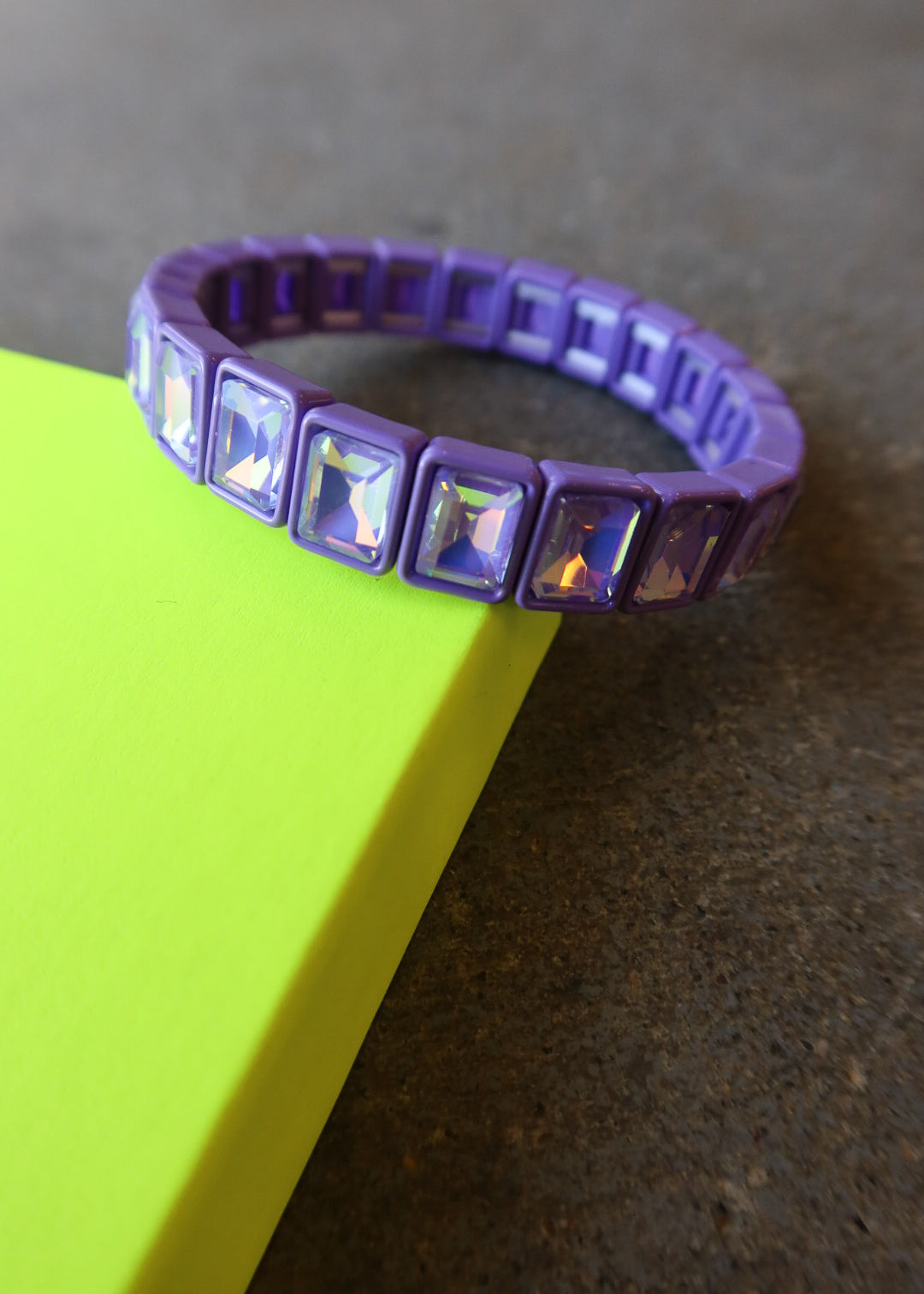 A purple silicone bracelet with rectangular iridescent rhinestones, resting on a bright yellow-green surface against a dark gray background