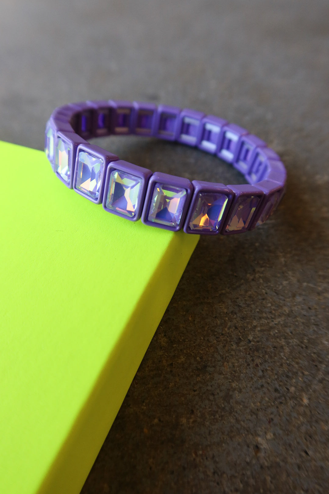 A purple silicone bracelet with rectangular iridescent rhinestones, resting on a bright yellow-green surface against a dark gray background