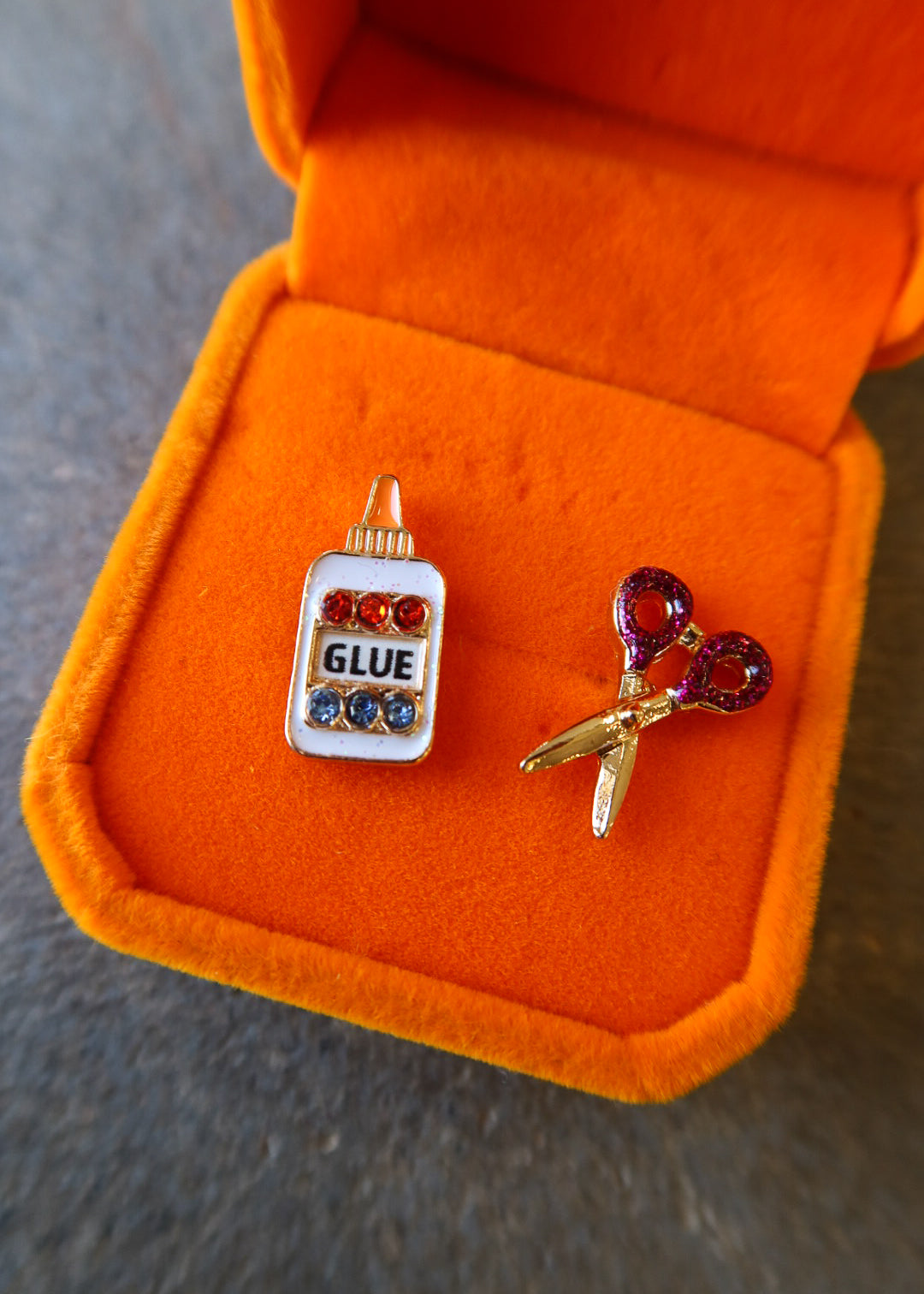 A pair of novelty earrings displayed in an open orange jewelry box on a gray surface. One earring is shaped like a glue bottle with red and blue rhinestones and the word 'GLUE' in black