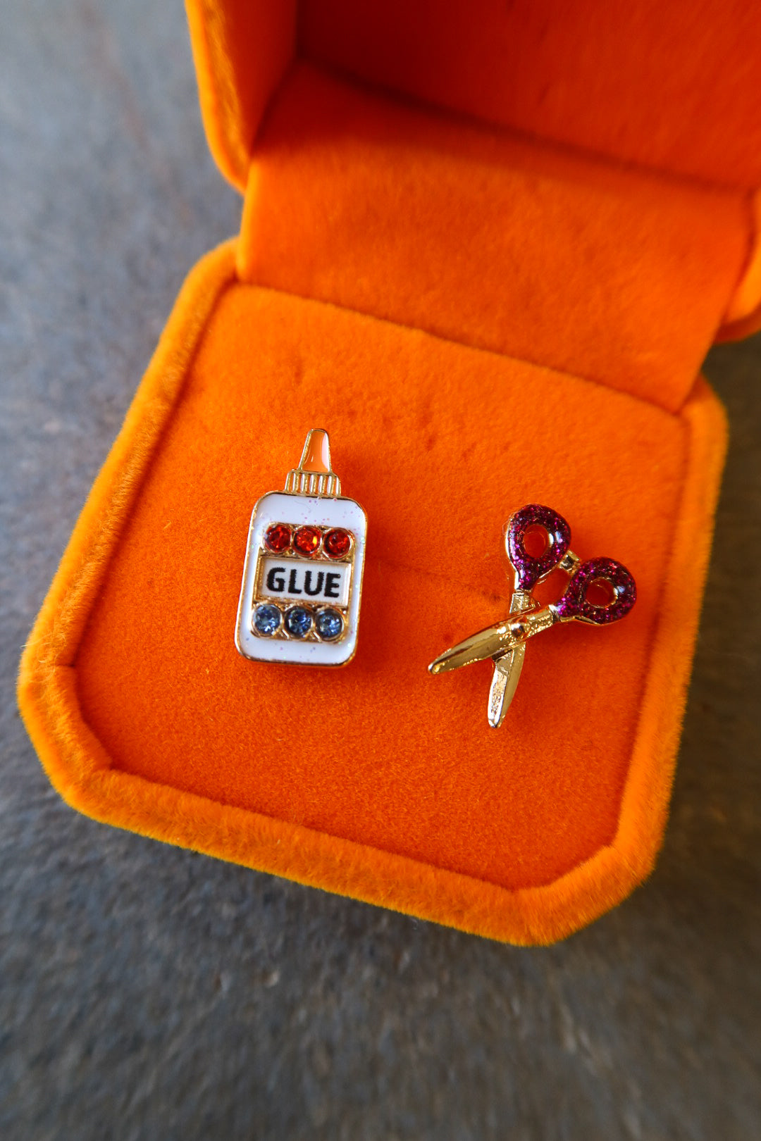 A pair of novelty earrings displayed in an open orange jewelry box on a gray surface. One earring is shaped like a glue bottle with red and blue rhinestones and the word 'GLUE' in black