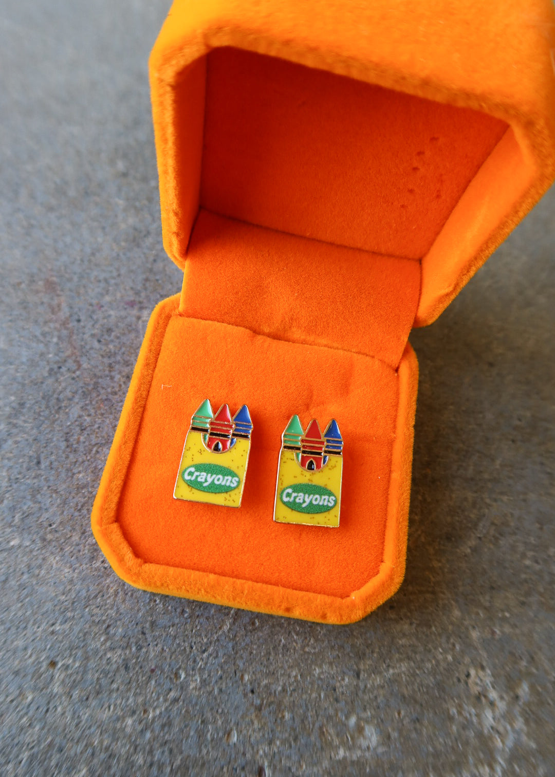 A pair of colorful Crayons enameled lapel pins displayed inside an open orange velvet jewelry box on a gray textured surface