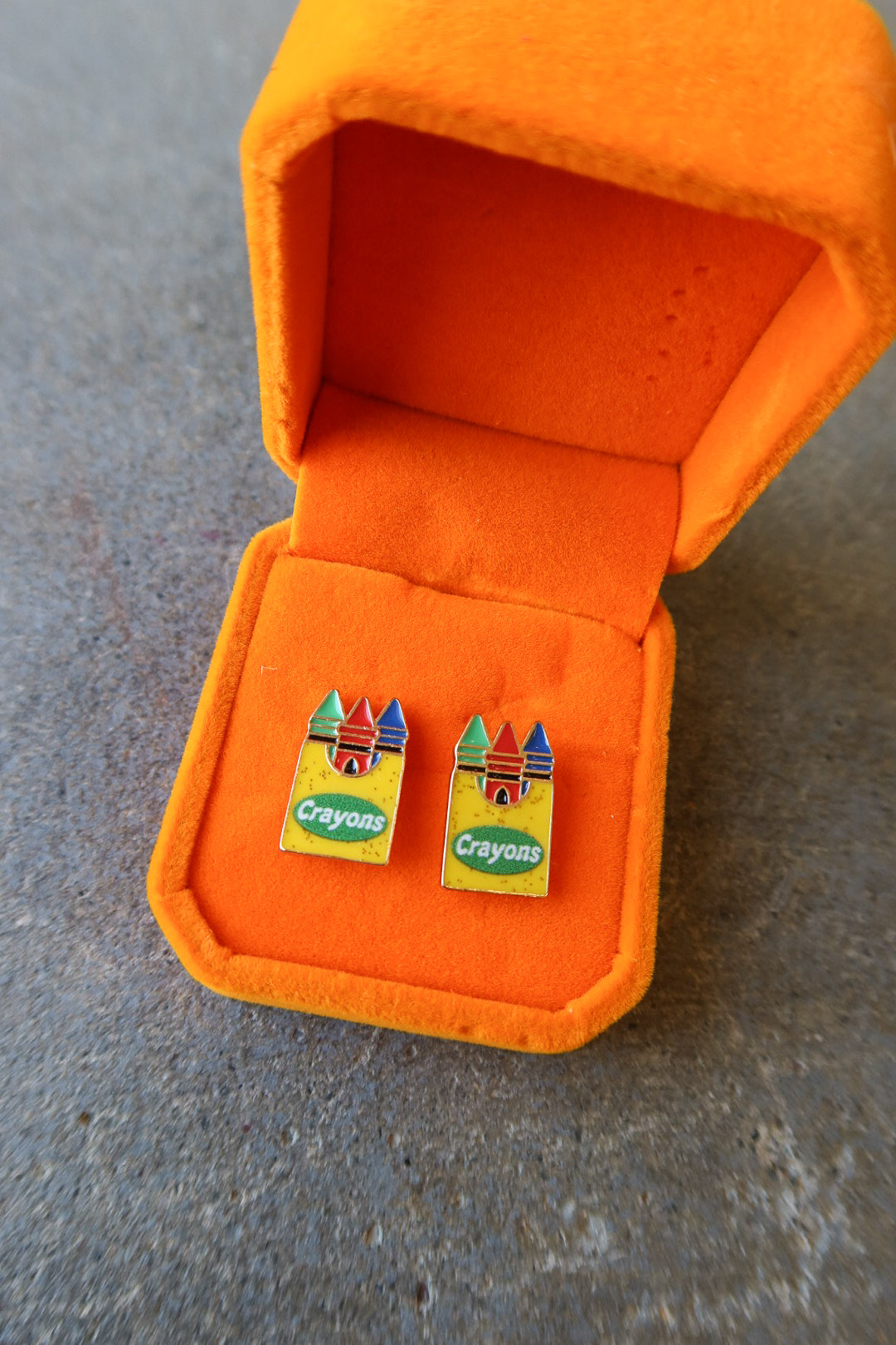 A pair of colorful Crayons enameled lapel pins displayed inside an open orange velvet jewelry box on a gray textured surface