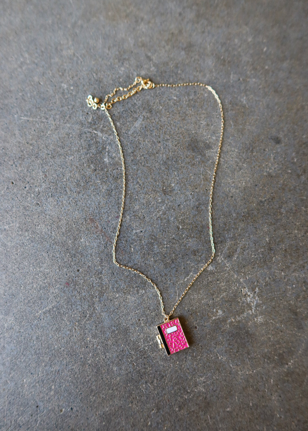 A delicate gold chain necklace featuring a small, vibrant pink textured pendant shaped like a book, displayed on a textured gray slate surface