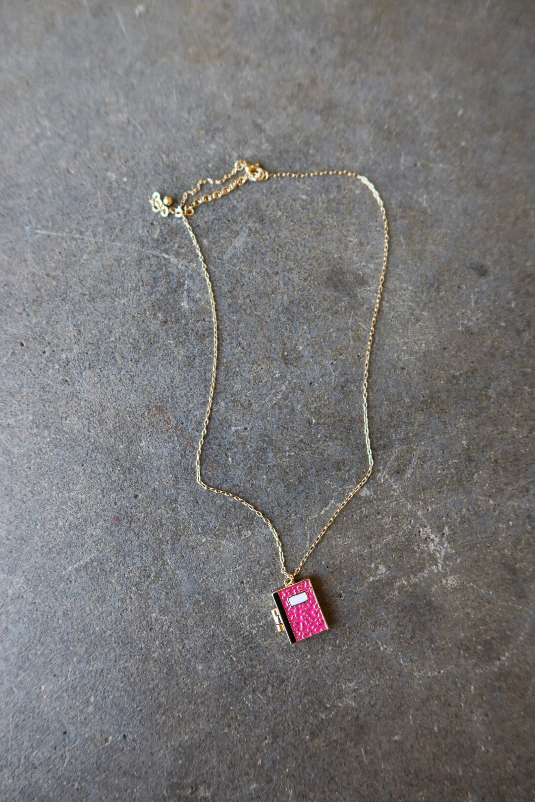 A delicate gold chain necklace featuring a small, vibrant pink textured pendant shaped like a book, displayed on a textured gray slate surface