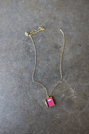 A delicate gold chain necklace featuring a small, vibrant pink textured pendant shaped like a book, displayed on a textured gray slate surface