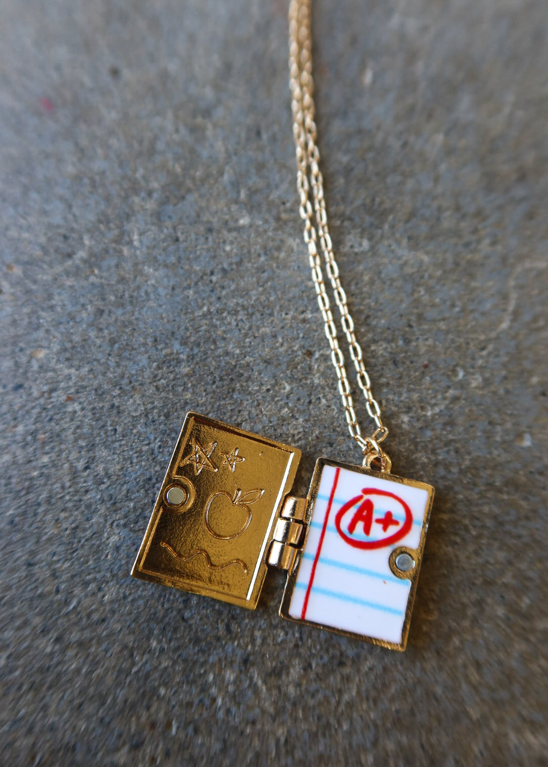 A gold-toned necklace with a rectangular locket pendant shaped like an open notebook. One side shows engraved symbols, and the other side has a lined paper design with a red circled 'A+'