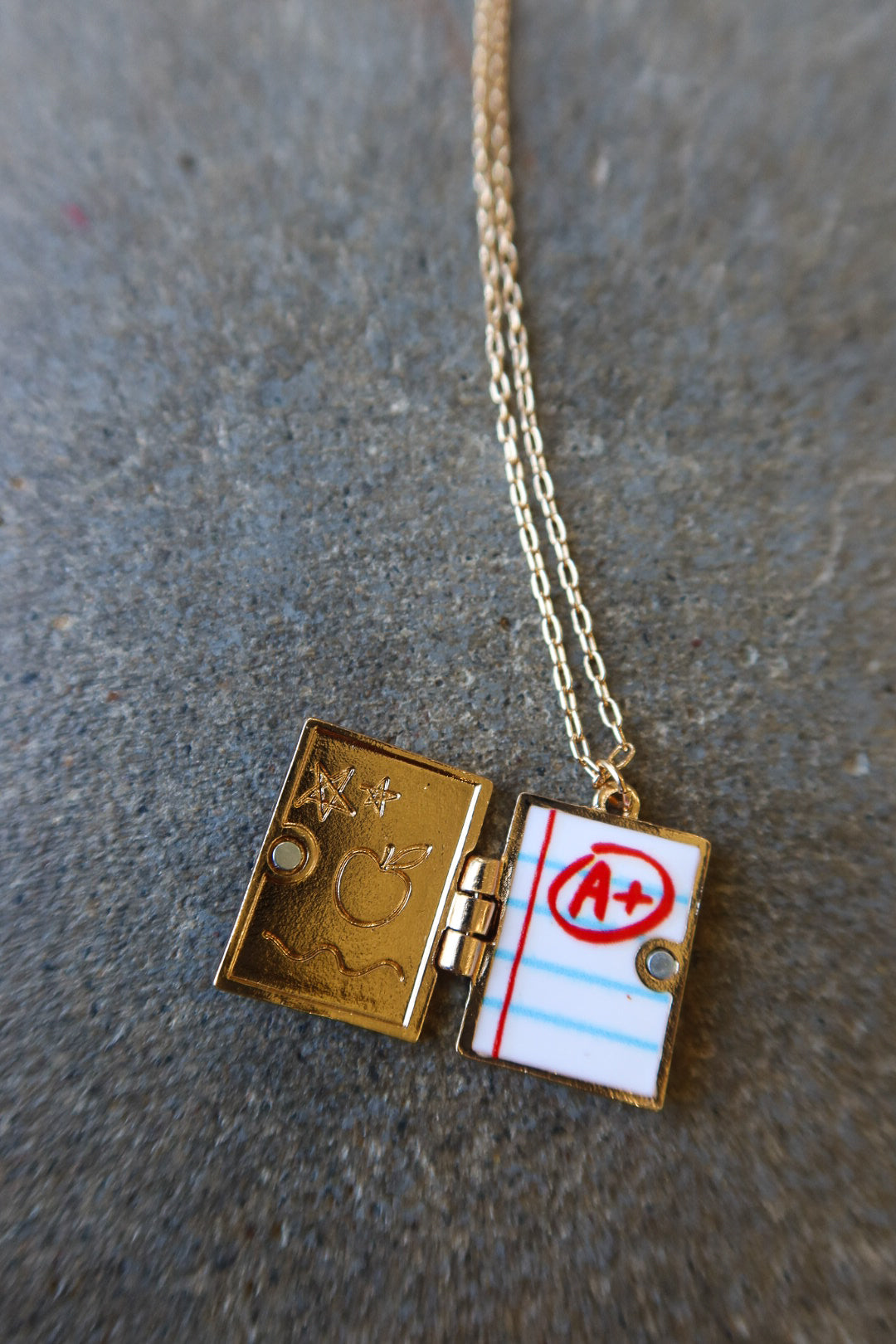 A gold-toned necklace with a rectangular locket pendant shaped like an open notebook. One side shows engraved symbols, and the other side has a lined paper design with a red circled 'A+'