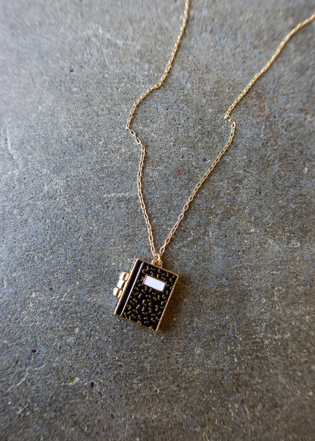A gold chain necklace with a small book-shaped pendant featuring a black speckled pattern and a white label, displayed on a textured gray stone surface