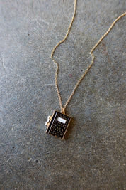A gold chain necklace with a small book-shaped pendant featuring a black speckled pattern and a white label, displayed on a textured gray stone surface