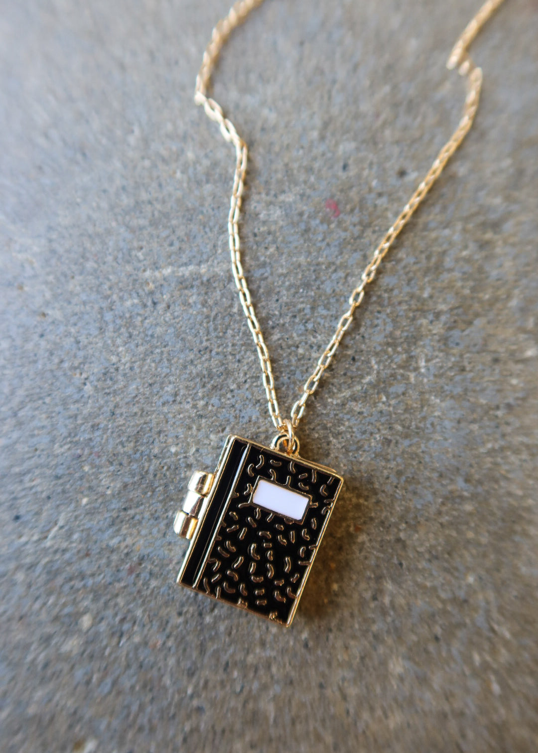 A gold chain necklace with a small book-shaped pendant featuring a textured black cover, gold edges, and a white rectangular detail on the front, displayed on a gray textured surface