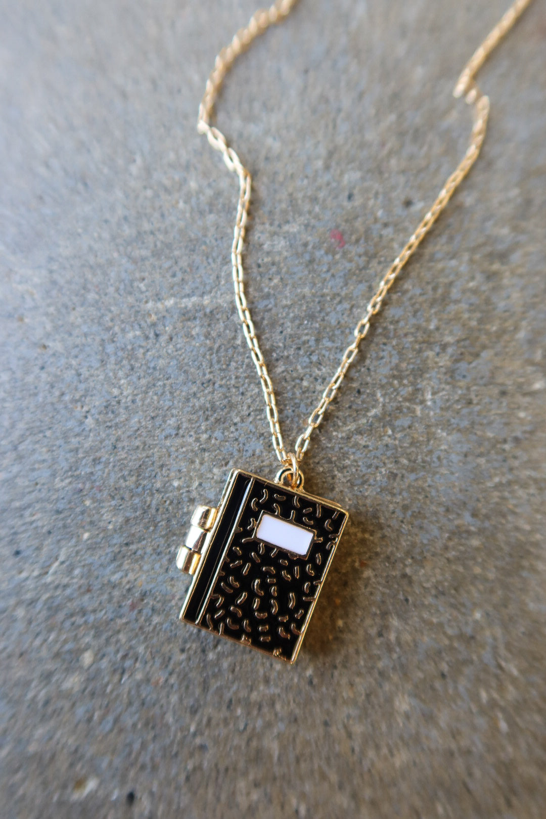 A gold chain necklace with a small book-shaped pendant featuring a textured black cover, gold edges, and a white rectangular detail on the front, displayed on a gray textured surface