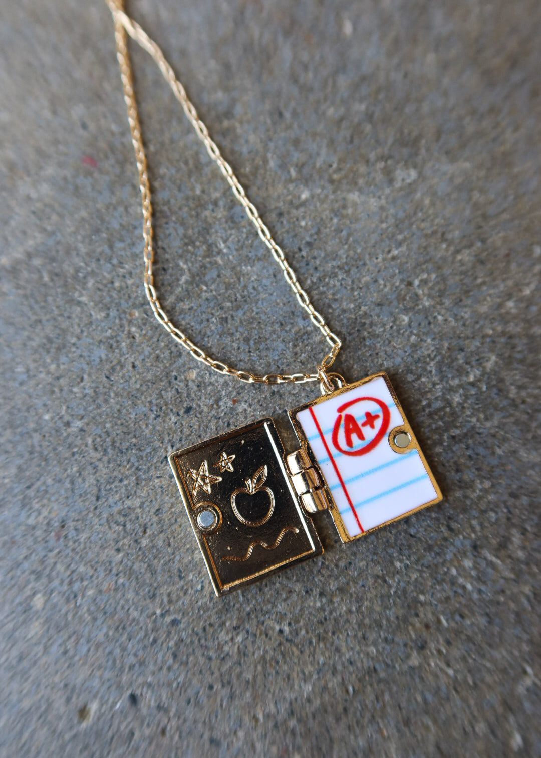 A gold chain necklace with a split locket pendant. One side of the locket is dark gold with an apple and stars motif, and the other side is white lined paper with red lines and a red circle over an 'A+