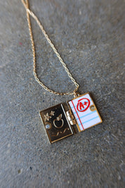 A gold chain necklace with a split locket pendant. One side of the locket is dark gold with an apple and stars motif, and the other side is white lined paper with red lines and a red circle over an 'A+