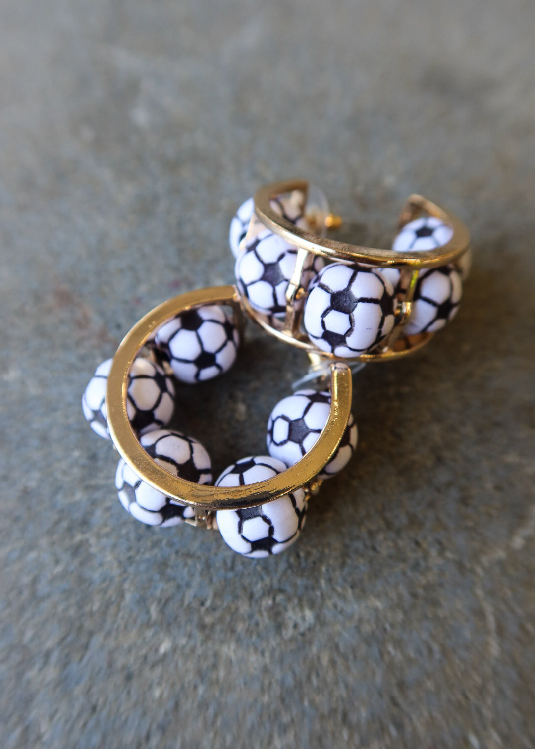 A pair of gold-tone cuff earrings decorated with white beads printed in a black soccer ball pattern, resting on a textured gray surface