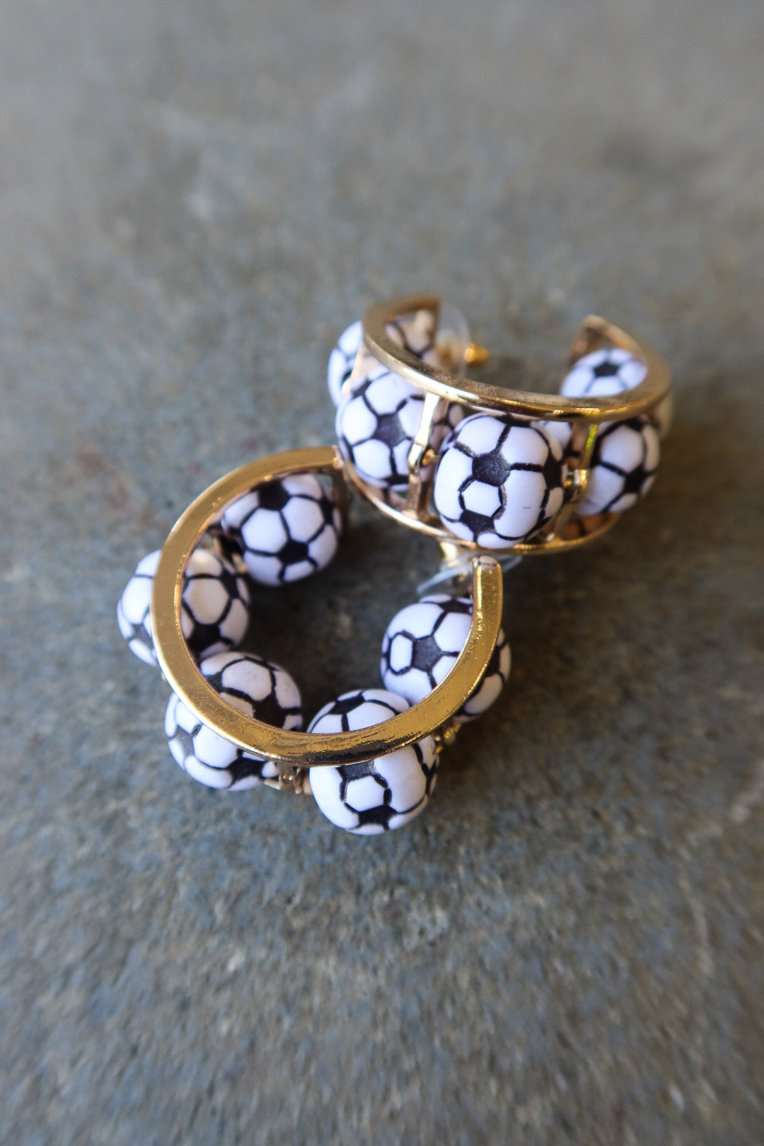 A pair of gold-tone cuff earrings decorated with white beads printed in a black soccer ball pattern, resting on a textured gray surface