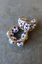 A pair of gold-tone cuff earrings decorated with white beads printed in a black soccer ball pattern, resting on a textured gray surface