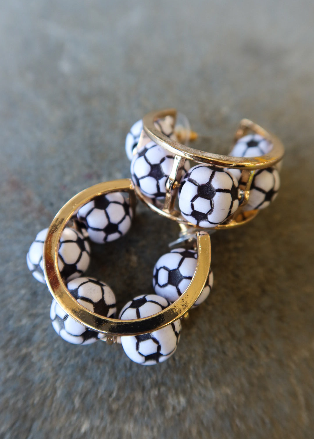 A pair of gold-tone hoop earrings decorated with white ceramic beads printed in a black soccer ball pattern, resting on a textured gray surface