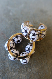 A pair of gold-tone hoop earrings decorated with white ceramic beads printed in a black soccer ball pattern, resting on a textured gray surface