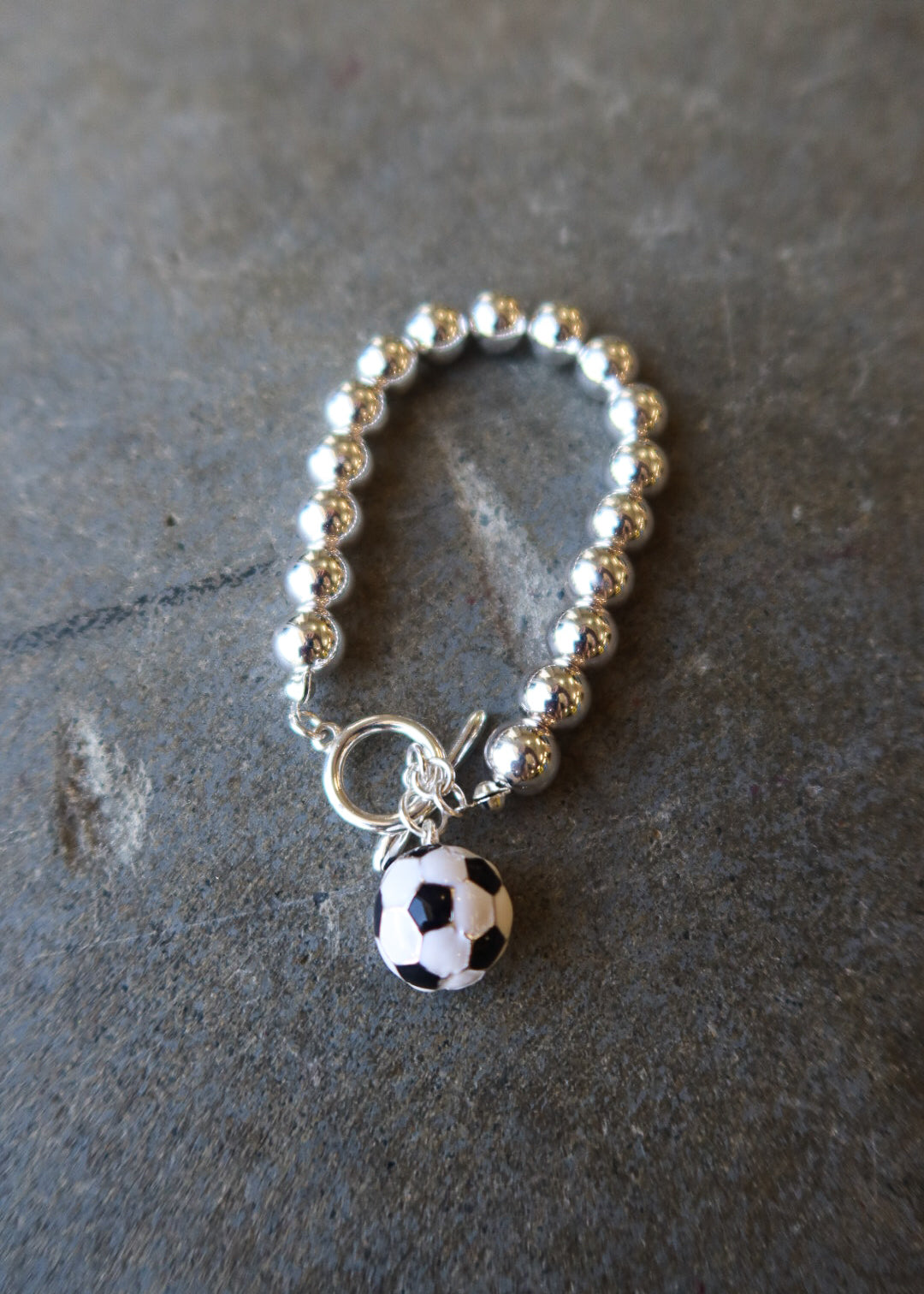 A silver beaded bracelet with a toggle clasp and a dangling soccer ball charm, displayed on a dark textured slate surface