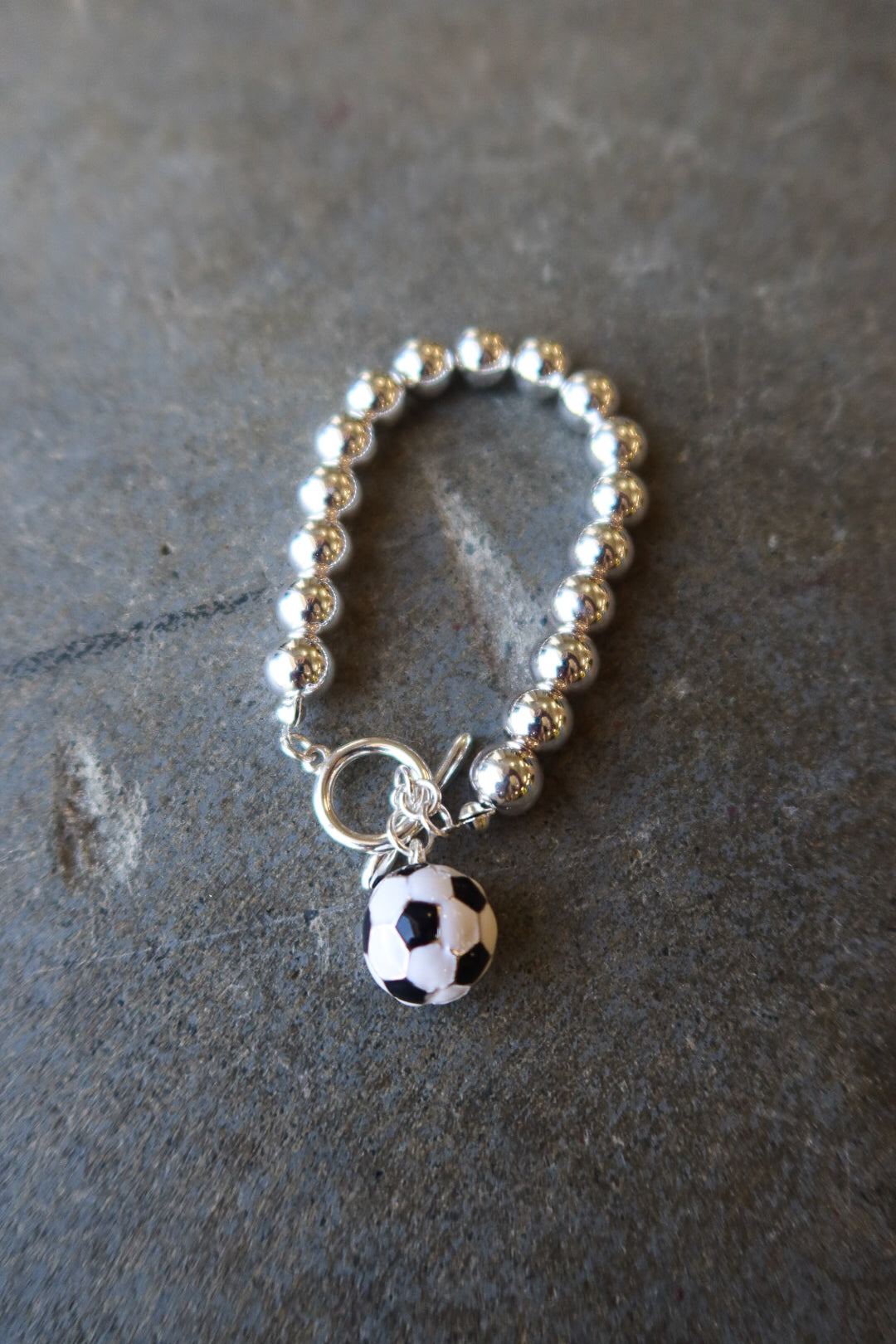 A silver beaded bracelet with a toggle clasp and a dangling soccer ball charm, displayed on a dark textured slate surface