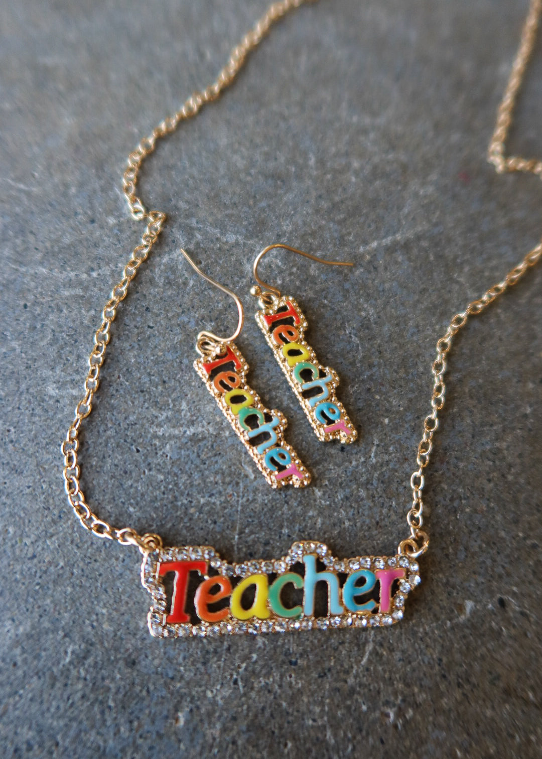 A gold-tone jewelry set featuring a necklace and matching drop earrings with the word 'Teacher' spelled out in colorful lettering, displayed on a textured gray surface