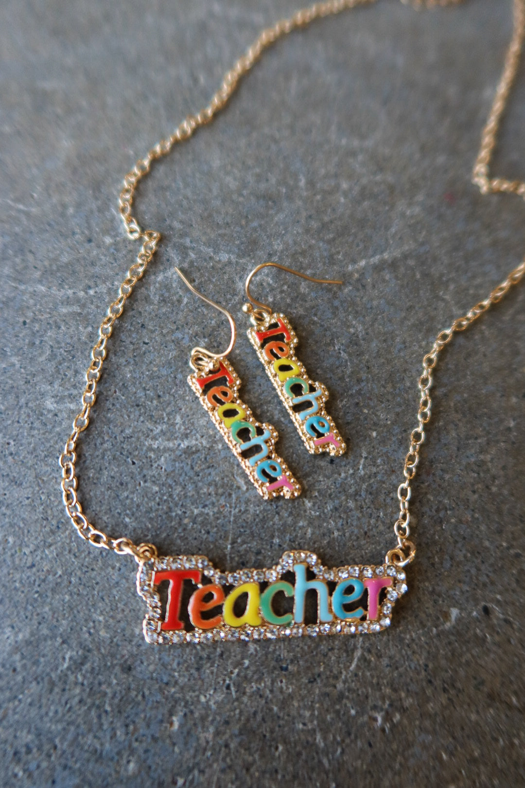 A gold-tone jewelry set featuring a necklace and matching drop earrings with the word 'Teacher' spelled out in colorful lettering, displayed on a textured gray surface