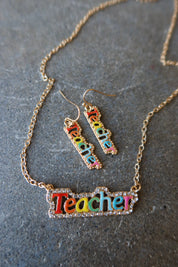 A gold-tone jewelry set featuring a necklace and matching drop earrings with the word 'Teacher' spelled out in colorful lettering, displayed on a textured gray surface