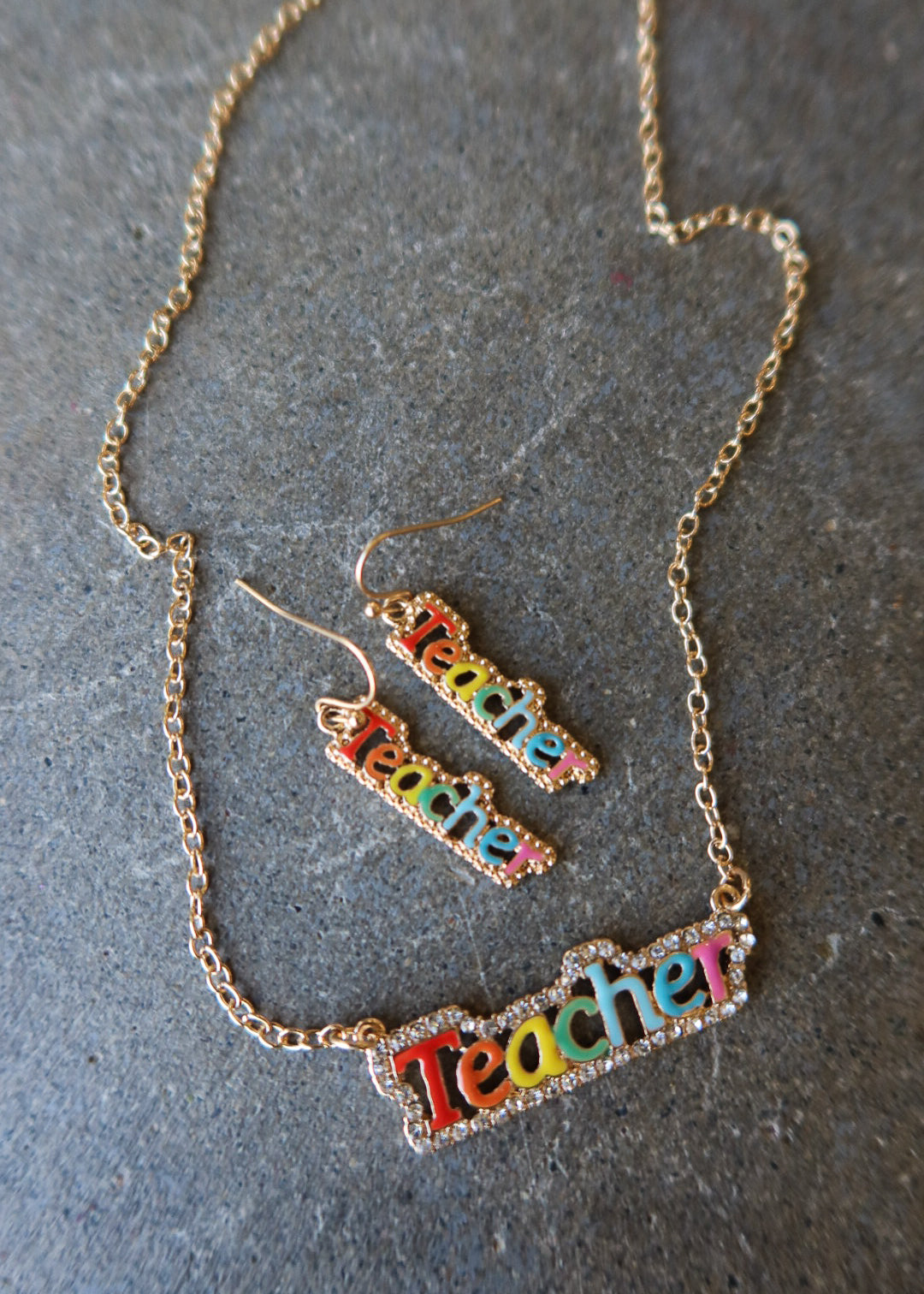 A gold-tone chain necklace and matching drop earrings with the word 'Teacher' spelled out in colorful lettering, displayed on a textured gray surface
