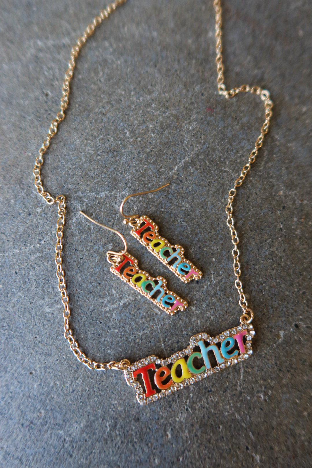 A gold-tone chain necklace and matching drop earrings with the word 'Teacher' spelled out in colorful lettering, displayed on a textured gray surface