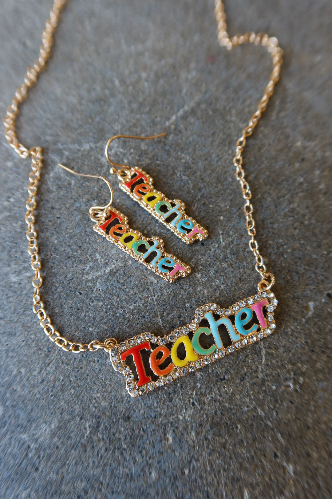 A gold-tone jewelry set featuring a rectangular pendant necklace and matching drop earrings, both decorated with the word "Teacher" in colorful raised letters outlined with small rhinestones, displayed on a textured gray surface
