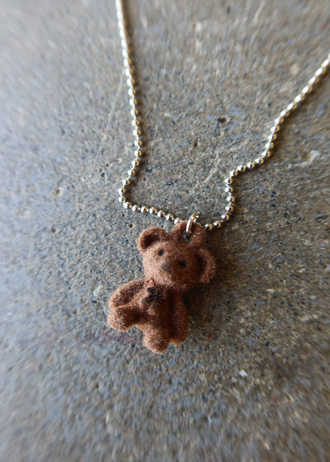 A small brown teddy bear pendant necklace on a silver ball chain, photographed on a textured gray surface