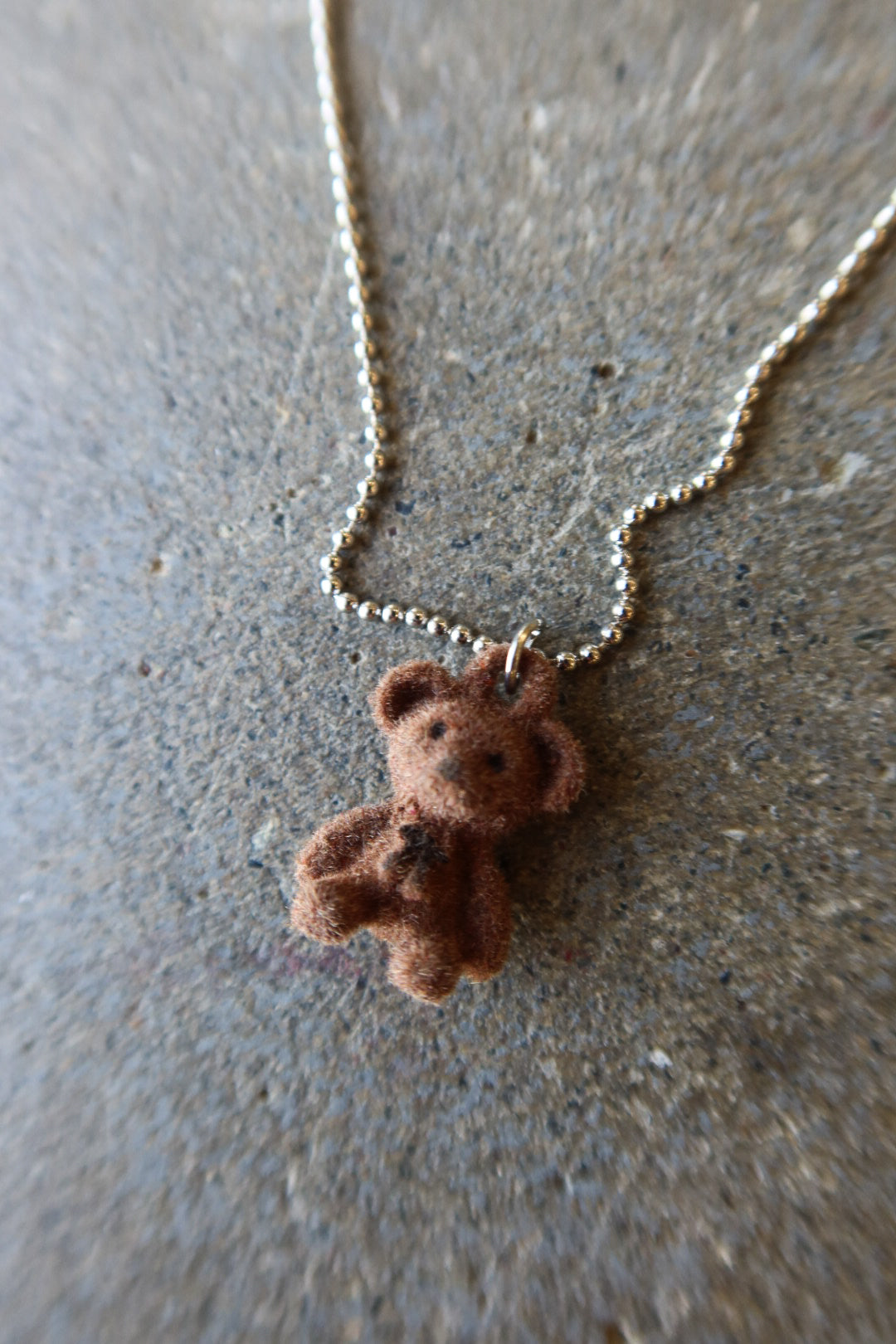 A small brown teddy bear pendant necklace on a silver ball chain, photographed on a textured gray surface
