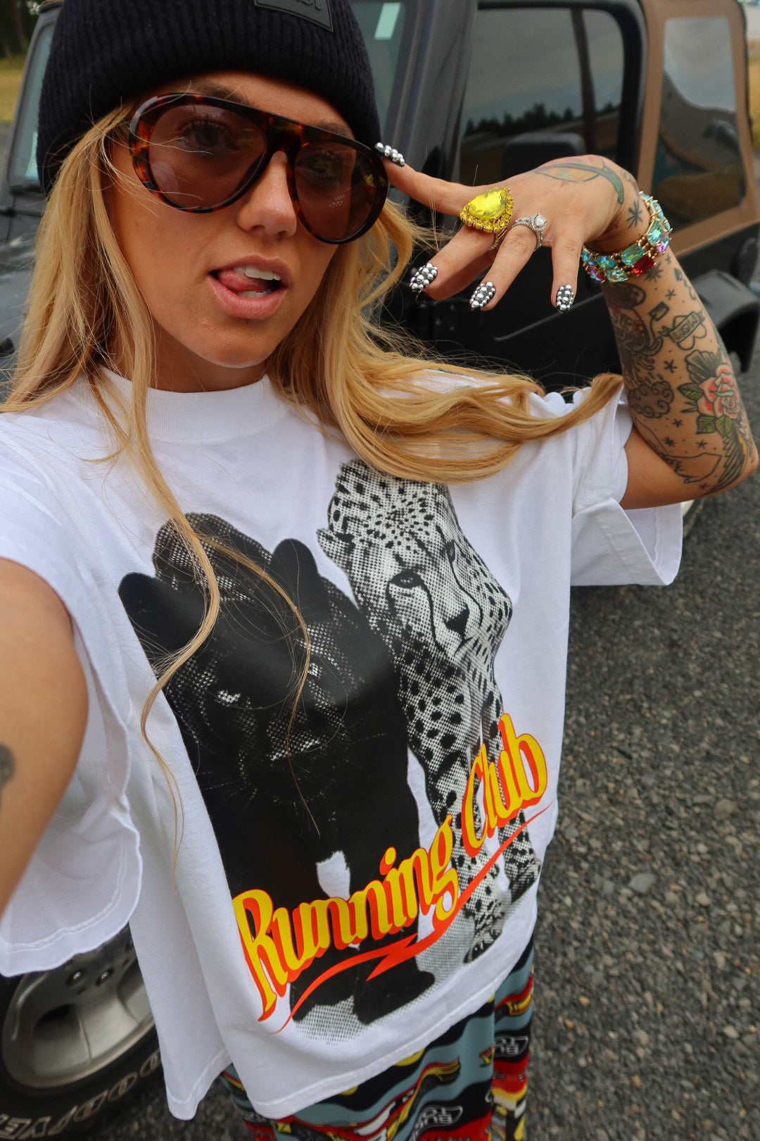 A woman wearing a white graphic t-shirt with a black leopard print and the words 'Running Club' in orange, paired with a black beanie, sunglasses, and bracelets, photographed outdoors in front of a vehicle