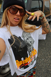 A woman wearing a white graphic t-shirt with a black leopard print and the words 'Running Club' in orange, paired with a black beanie, sunglasses, and bracelets, photographed outdoors in front of a vehicle